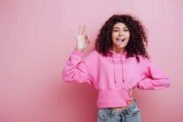 Alegre bi-racial menina saindo língua e mostrando ok gesto no fundo rosa — Fotografia de Stock