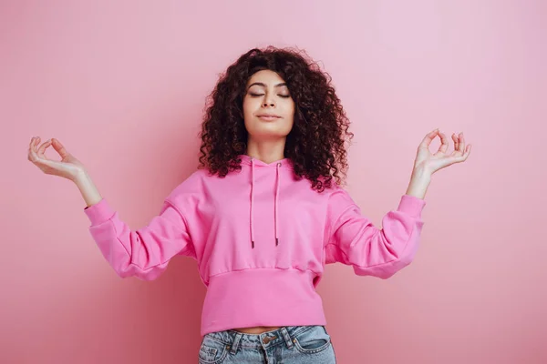 Pretty bi-racial girl meditating with closed eyes while standing on pink background — Stock Photo
