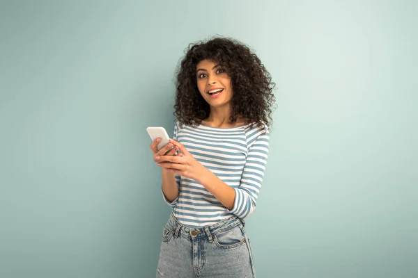Feliz bi-racial chica charlando en el teléfono inteligente mientras mira a la cámara en el fondo gris - foto de stock