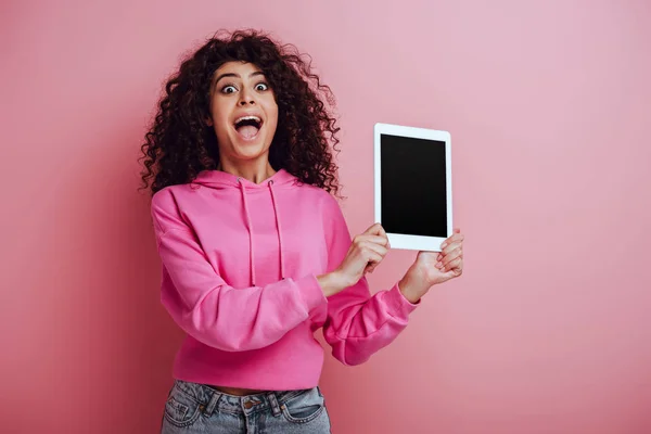 Conmocionada chica bi-racial mostrando tableta digital con pantalla en blanco sobre fondo rosa — Stock Photo