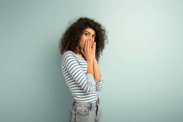 Scared bi-racial girl covering face with hands while looking at camera on grey background — Stock Photo