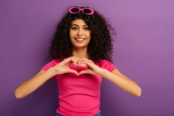 Feliz bi-racial chica mostrando el signo del corazón con las manos mientras mira a la cámara en el fondo púrpura - foto de stock