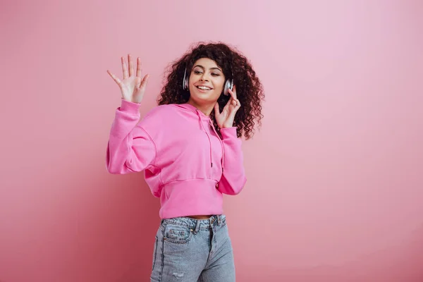 Cheerful bi-racial girl waving hand while listening music in wireless headphones on pink background — Stock Photo