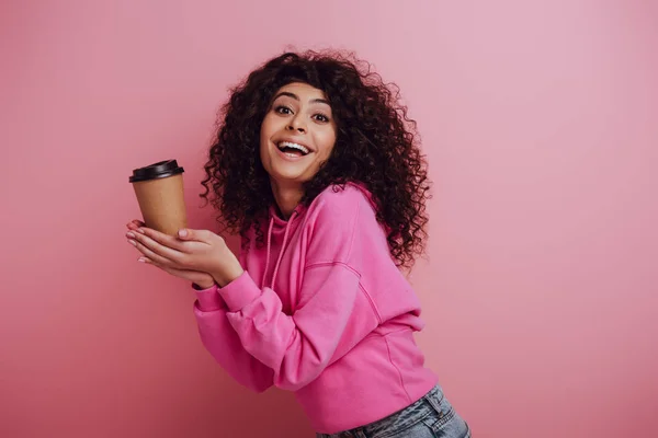 Happy bi-racial girl smiling at camera while holding coffee to go on pink background — Stock Photo