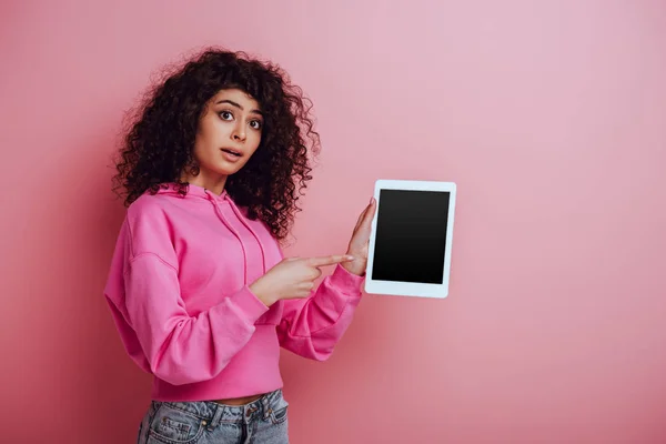 Приваблива бі-расова дівчина, що вказує пальцем на цифровий планшет з порожнім екраном на рожевому фоні — стокове фото