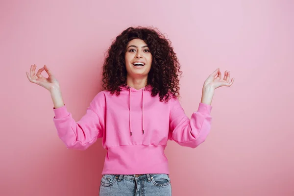 Cheerful bi-racial girl looking at camera while standing in meditation pose on pink background — Stock Photo