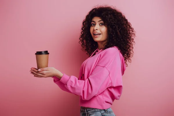 Pretty bi-racial girl holding coffee to go and smiling at camera on pink background — Stock Photo