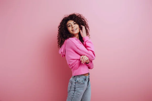 Flirty, smiling bi-racial girl touching hair while smiling at camera on pink background — Stock Photo