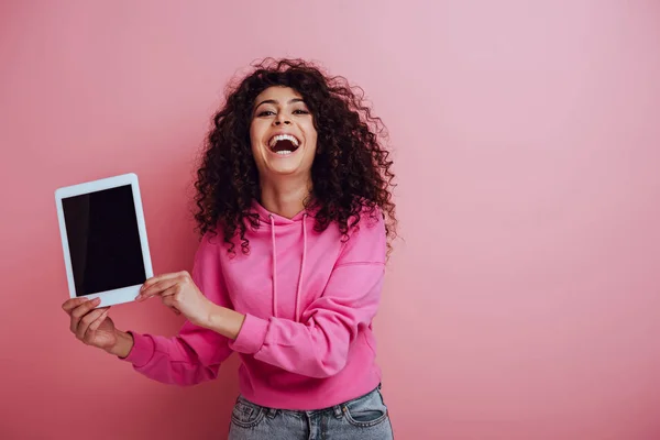 Смеющаяся двурасовая девушка показывает цифровой планшет с чистым экраном на розовом фоне — стоковое фото