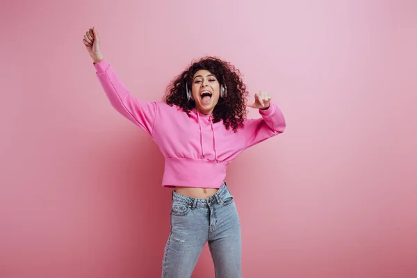 Aufgeregtes gemischtes Rassenmädchen tanzt und singt, während es Musik in drahtlosen Kopfhörern auf rosa Hintergrund hört — Stockfoto