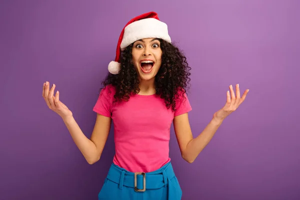 Amazed bi-racial girl in santa hat looking at camera while standing with open arms on purple background — Stock Photo