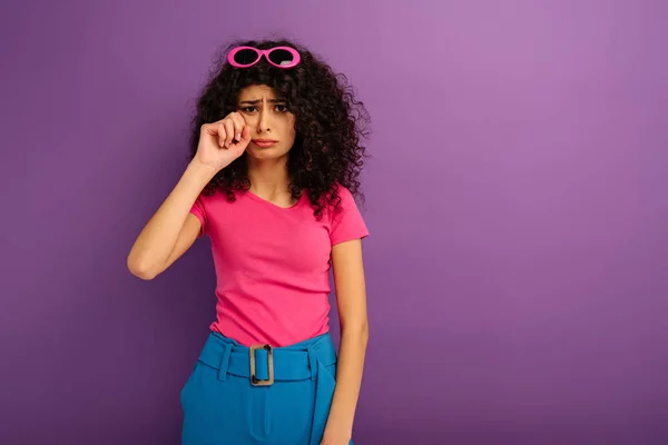 Обиженная двурасовая девушка, вытирающая слезы рукой, плача на фиолетовом фоне — стоковое фото