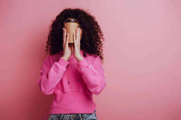 Fille de race mixte en sweat à capuche rose visage obscurcissant avec tasse en papier sur fond rose — Photo de stock