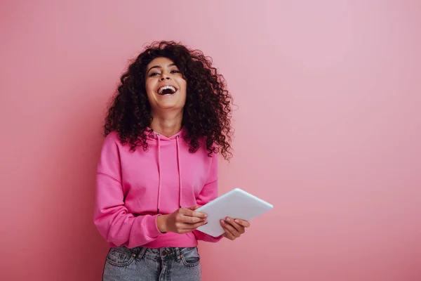 Щаслива бі-расова жінка сміється з камери, тримаючи цифровий планшет на рожевому фоні — стокове фото