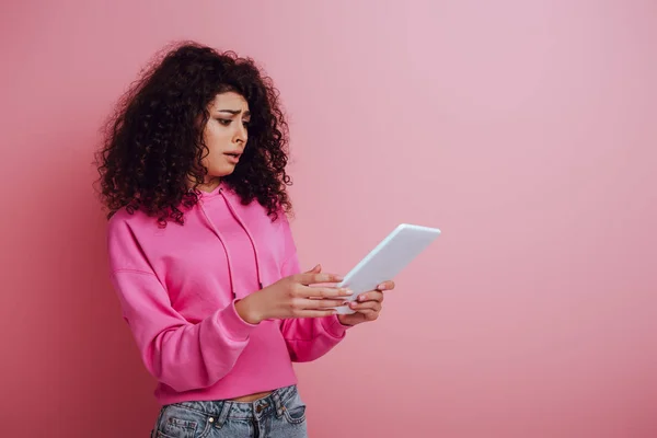 Unzufriedenes zweirassiges Mädchen, das auf ein digitales Tablet auf rosa Hintergrund blickt — Stockfoto