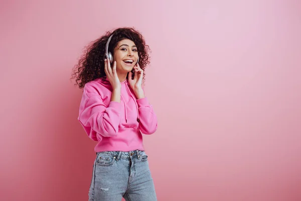 Glückliches zweirassiges Mädchen, das in die Kamera lächelt, während es Musik in drahtlosen Kopfhörern auf rosa Hintergrund hört — Stock Photo