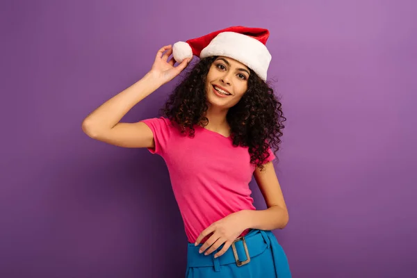 Feliz bi-racial menina tocando santa chapéu e sorrindo para a câmera no fundo roxo — Fotografia de Stock