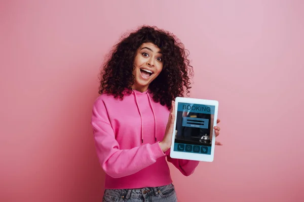 Aufgeregtes zweirassiges Mädchen zeigt digitales Tablet mit Buchungs-App auf rosa Hintergrund — Stockfoto