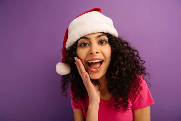 Aufgeregtes bi-rassisches Mädchen mit Weihnachtsmann-Hut, das vor der Kamera auf lila Hintergrund schreit — Stockfoto