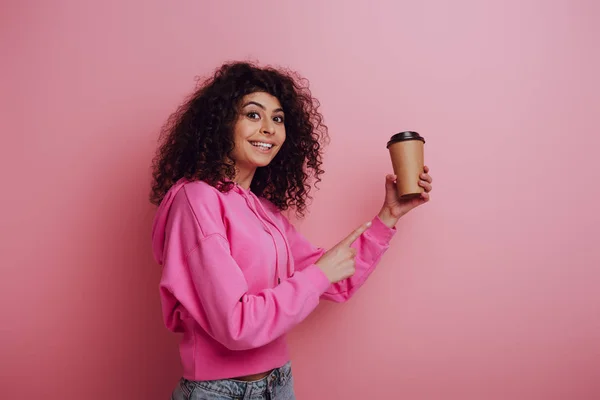 Glückliches zweirassiges Mädchen, das mit dem Finger auf Coffee to go zeigt, während es auf rosa Hintergrund in die Kamera lächelt — Stockfoto