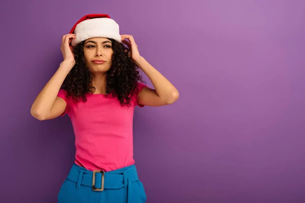 Скептична бі-расова дівчина торкається капелюха Санти і дивиться на фіолетовий фон — стокове фото