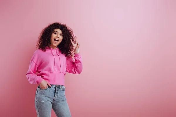 Alegre bi-racial menina mostrando vitória gesto enquanto segurando mão no bolso no fundo rosa — Fotografia de Stock