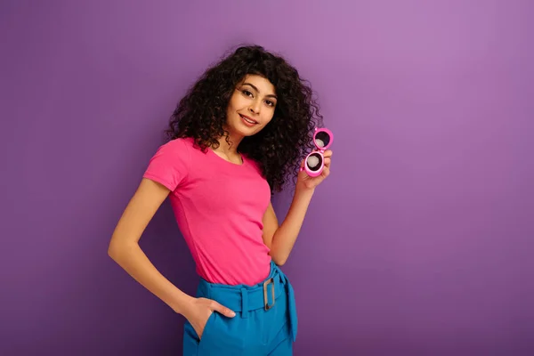 Attractive bi-racial girl holding sunglasses while smiling at camera on purple background — Stock Photo