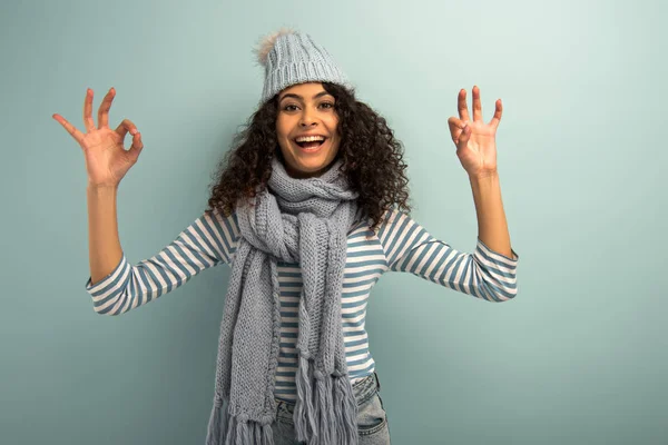 Happy bi-racial girl in warm hat and scarf showing okay signs while looking at camera on grey background — Stock Photo