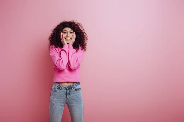 Щаслива бі-расова дівчина посміхається на камеру, торкаючись обличчя на рожевому фоні — стокове фото