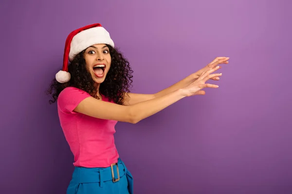 Excited bi-racial girl in santa hat showing frightening gesture with hands on purple background — Stock Photo