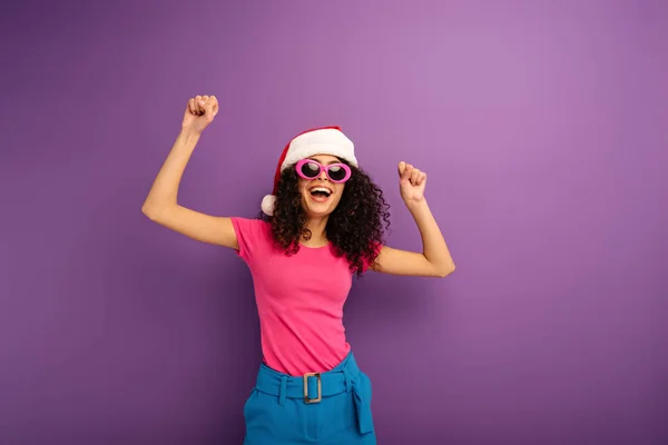 Fröhliches zweirassiges Mädchen mit Weihnachtsmütze und Sonnenbrille, das auf lila Hintergrund tanzt — Stockfoto