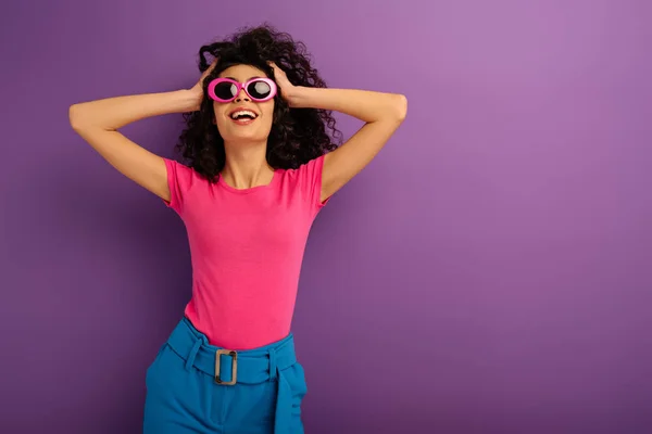 Счастливая двурасовая девушка в солнечных очках трогает голову и улыбается на фиолетовом фоне — стоковое фото
