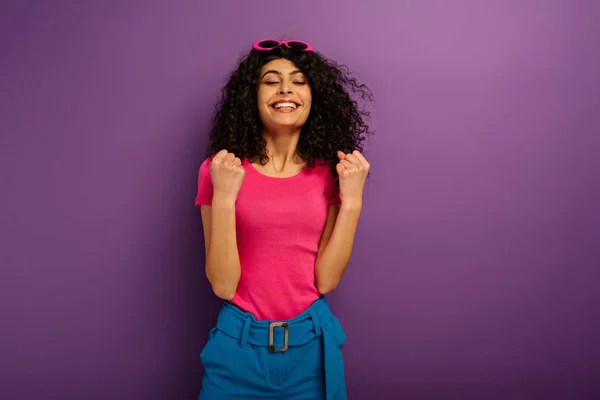 Excited bi-racial girl showing winner gesture while standing with closed eyes on purple background — Stock Photo