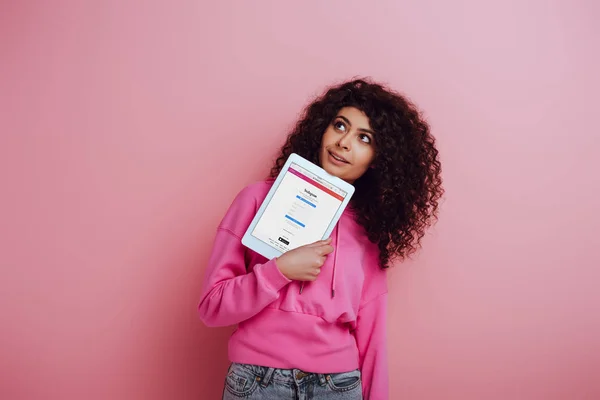 KYIV, UCRANIA - 26 de noviembre de 2019: chica bi-racial soñadora mirando hacia otro lado mientras muestra la tableta digital con la aplicación Instagram sobre fondo rosa - foto de stock
