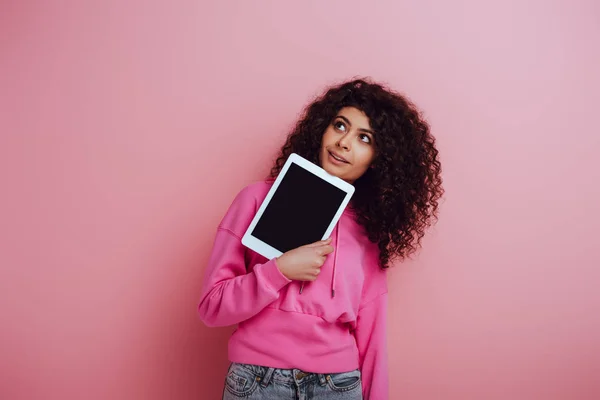 Мечтательная би-расовая девушка смотрит в сторону, показывая цифровой планшет с чистым экраном на розовом фоне — стоковое фото