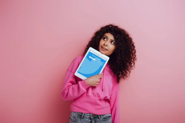 KYIV, UKRAINE - NOVEMBER 26, 2019: dreamy bi-racial girl looking away while showing digital tablet with Twitter app on pink background — Stock Photo