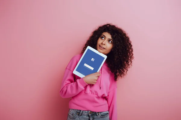 KYIV, UCRANIA - 26 de noviembre de 2019: chica bi-racial soñadora mirando hacia otro lado mientras muestra la tableta digital con la aplicación de Facebook sobre fondo rosa - foto de stock