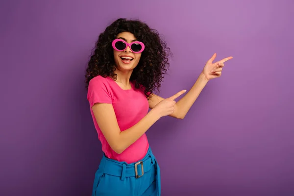 Happy bi-racial girl in sunglasses looking at camera while pointing with fingers on purple background — Stock Photo