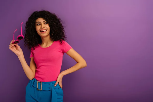 Cheerful bi-racial girl holding sunglasses while looking away on purple background — Stock Photo