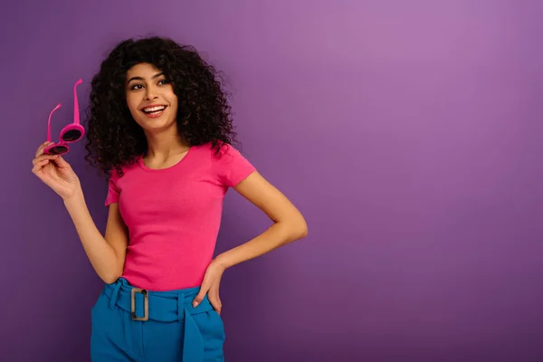 Joyeuse fille bi-raciale tenant des lunettes de soleil tout en regardant loin sur fond violet — Photo de stock