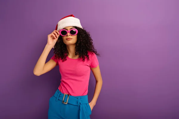 Стильная смешанная гонка девушка в шляпе Санты касаясь очки, держа руку в кармане на фиолетовом фоне — стоковое фото