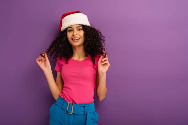 Привлекательная девушка смешанной расы в шапке Санты касаясь волос, улыбаясь в камеру на фиолетовом фоне — стоковое фото