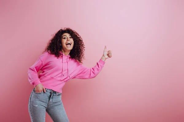 Feliz bi-racial menina mostrando polegar para cima enquanto segurando mão no bolso no fundo rosa — Fotografia de Stock