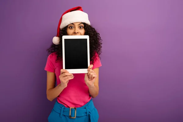 Chica de raza mixta positiva en sombrero de santa mirando a la cámara mientras que muestra tableta digital con pantalla en blanco sobre fondo púrpura - foto de stock
