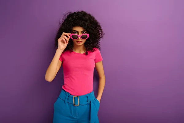 Уверенная двурасовая девушка касается солнцезащитных очков, глядя на камеру на фиолетовом фоне — стоковое фото