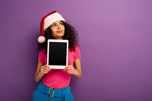 Fille de race mixte rêveuse dans le chapeau santa détournant les yeux tout en montrant tablette numérique avec écran blanc sur fond violet — Photo de stock