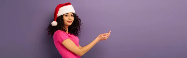 Plano panorámico de chica de raza mixta sonriente en sombrero de santa de pie con las manos dobladas sobre fondo púrpura - foto de stock
