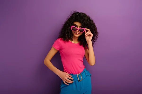 Привлекательная двурасовая девушка касается солнцезащитных очков, улыбаясь в камеру на фиолетовом фоне — стоковое фото