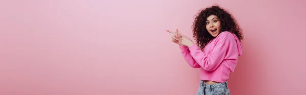 Панорамный снимок шокированной двурасовой девушки, смотрящей в камеру, указывая пальцами на розовый фон — стоковое фото