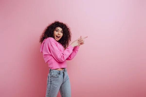 Шокована бі-расова дівчина дивиться на камеру, вказуючи пальцями на рожевий фон — стокове фото
